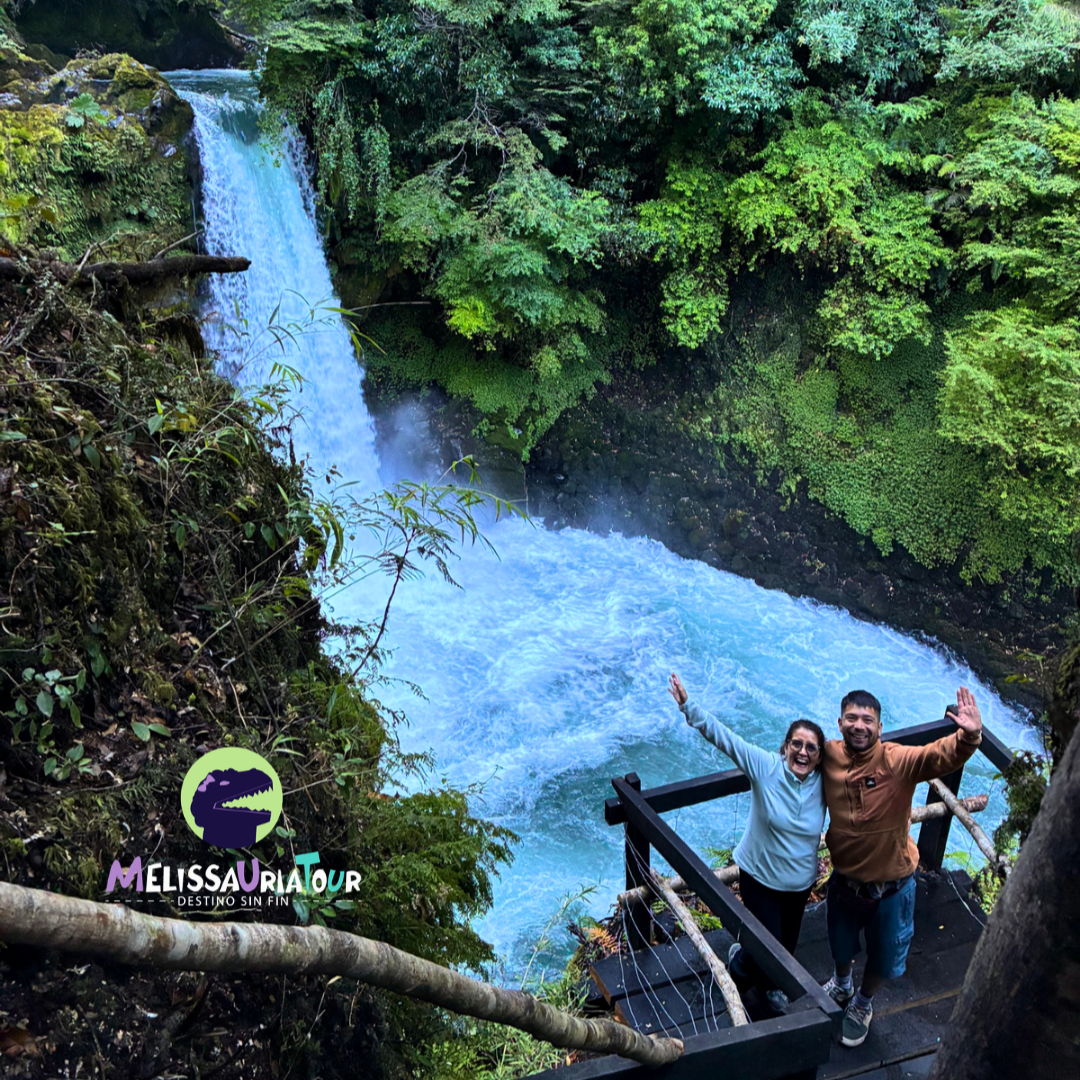 Turistas visitan un salto de agua de color turquesa desde un mirador en medio del bosque nativo.