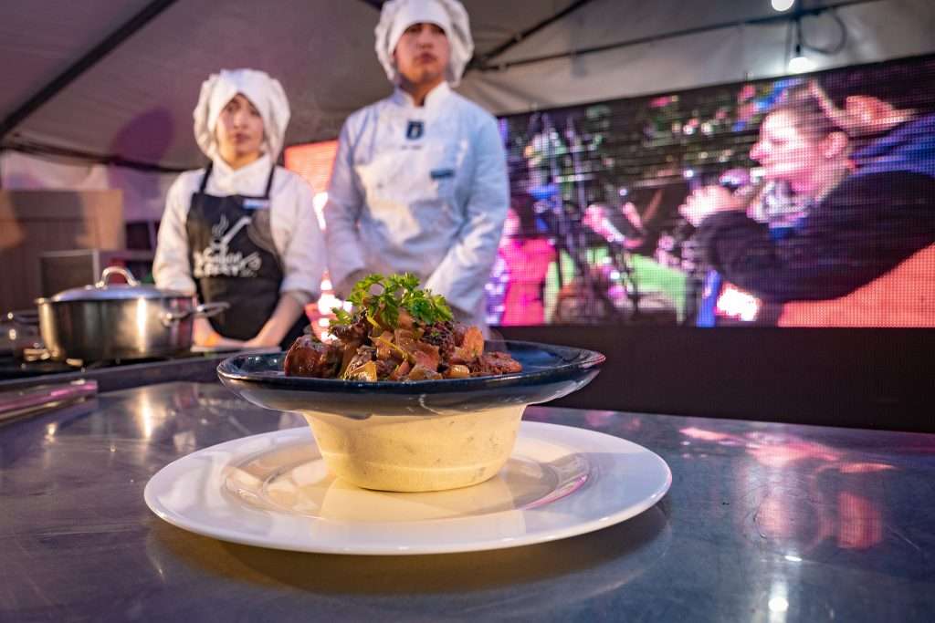 Plato de comida típica servido en un bol de greda sobre un escenario gastronómico, con chefs al fondo durante una demostración culinaria en Sabor Fest Panguipulli.