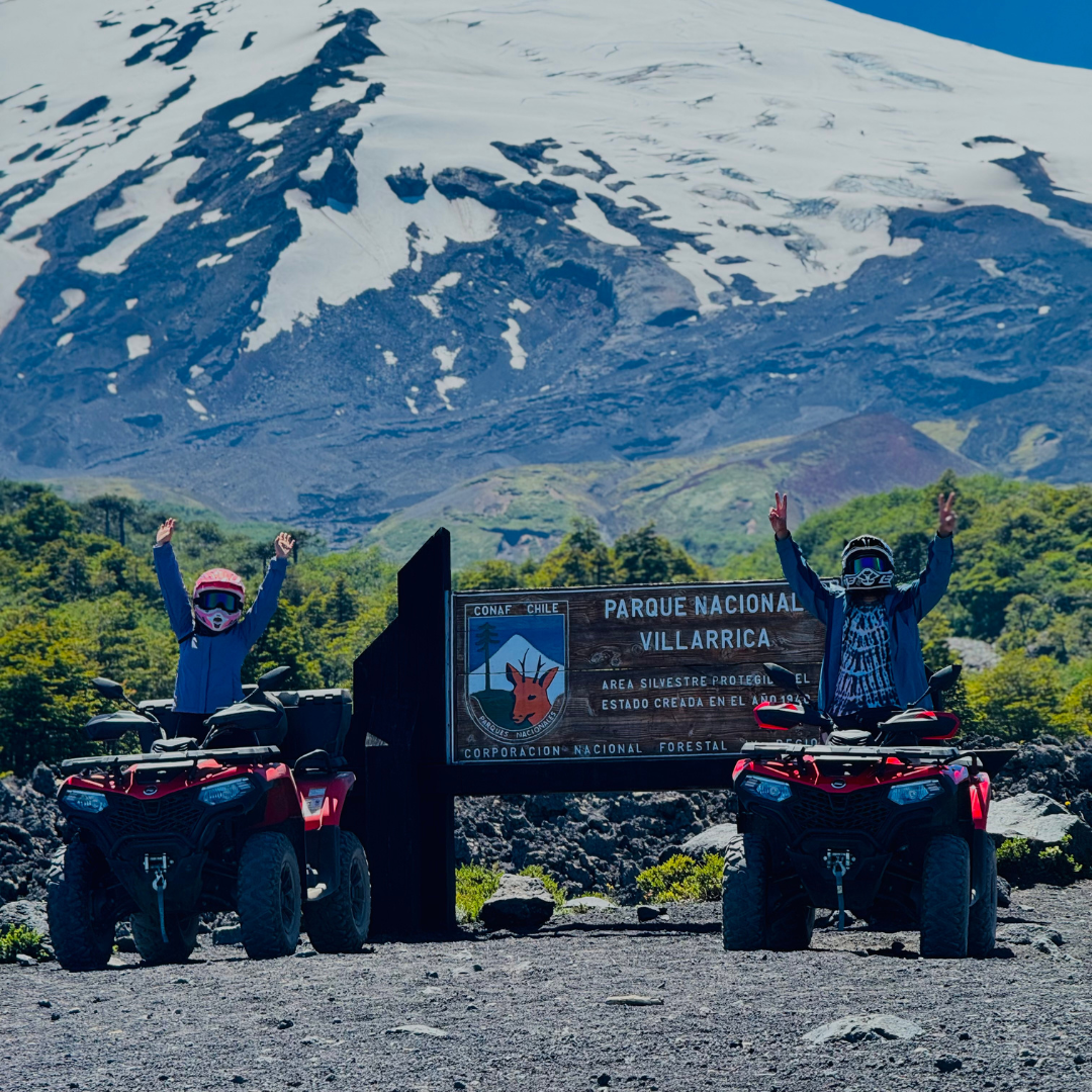 Personas en cuatrimotos junto al letrero del Parque Nacional Villarrica con el volcán nevado de fondo.